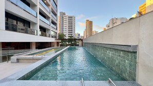 Une piscine dont le bord est revêtu de carreaux, située à côté d’un immeuble doté de balcons, sous un ciel dégagé.