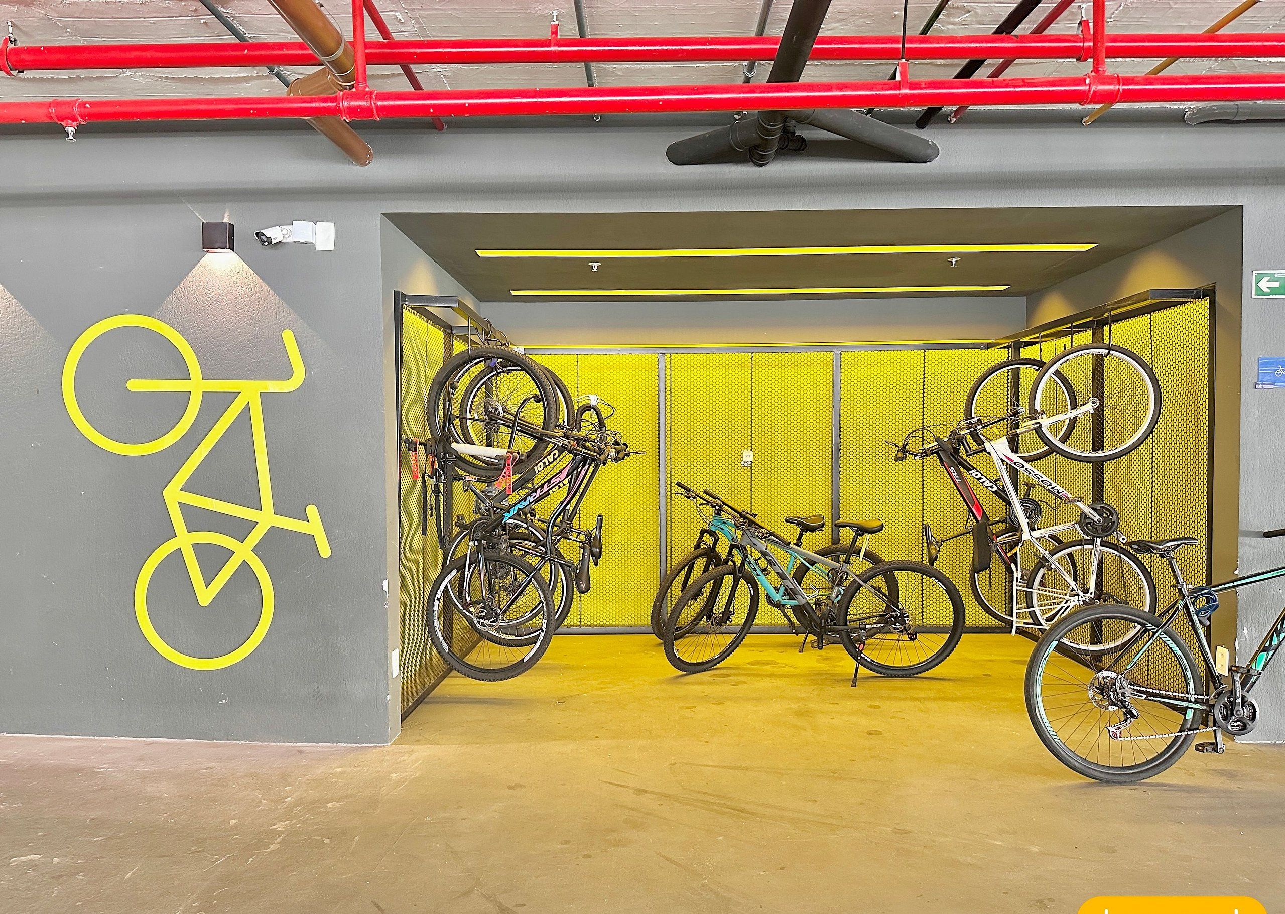 A bicycle parking area with multiple bikes, a yellow wall, and a red pipe overhead.