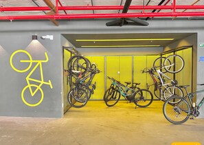 A bicycle parking area with multiple bikes, a yellow wall, and a red pipe overhead. - Jsmart Dom (Fortaleza)