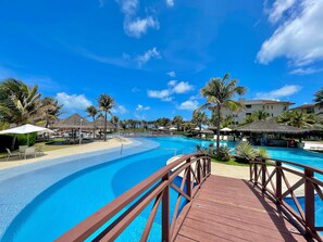 Une piscine d’hôtel avec une terrasse en bois, des palmiers et des parasols en chaume.
