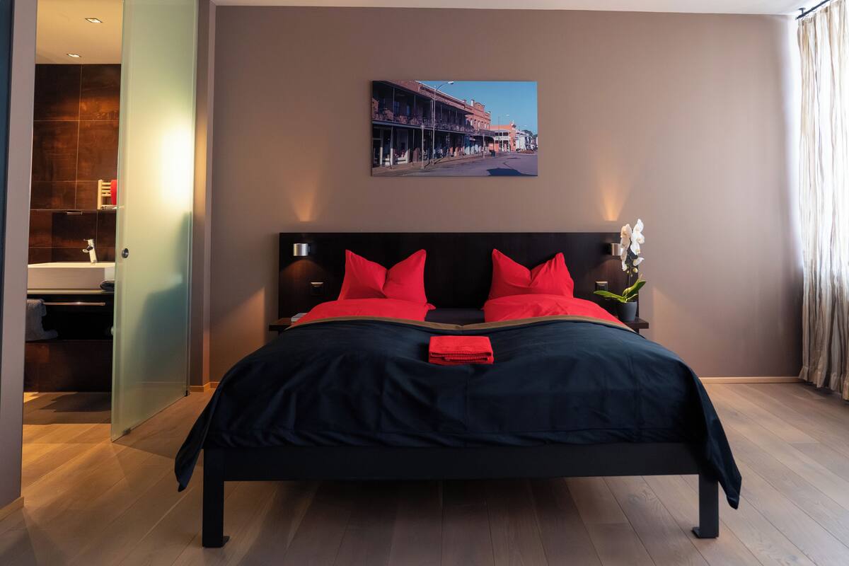 A modern bedroom with a large bed, a nightstand, a potted plant, and a cityscape painting on the wall.