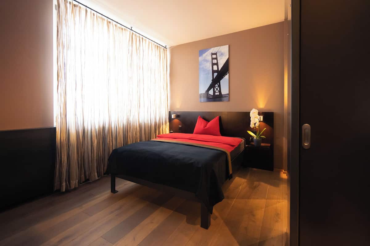 A hotel room with a bed, a nightstand, a lamp, a painting of the Golden Gate Bridge, and a window with curtains.