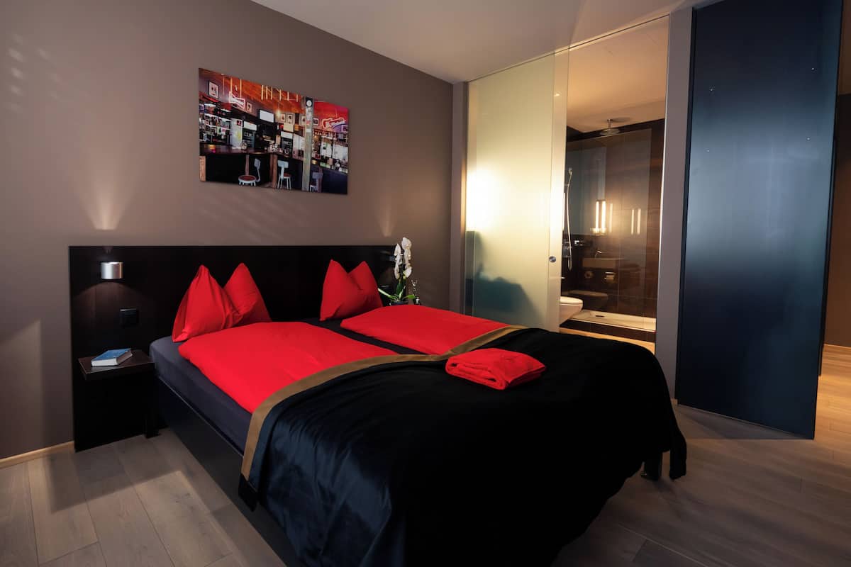 A modern hotel room with a large bed, a nightstand, a wall-mounted TV, and a bathroom visible through a glass door.