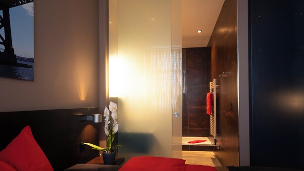 Ein modernes Hotelzimmer mit Bett, roten Kissen, einer Topfpflanze und Orchidee, einer Duschkabine und einem dunkelholzigen Kleiderschrank.