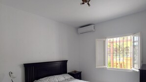 Une chambre à coucher comprenant un lit, une table de chevet, un bureau, une fenêtre avec des barreaux et un appareil de climatisation.
