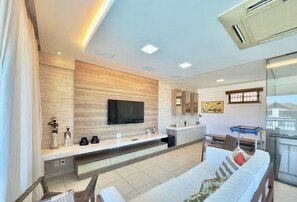 Quarto de hotel moderno com televisão de tela plana fixada em parede de madeira, sofá com almofadas, pequena mesa de jantar e varanda com vista.