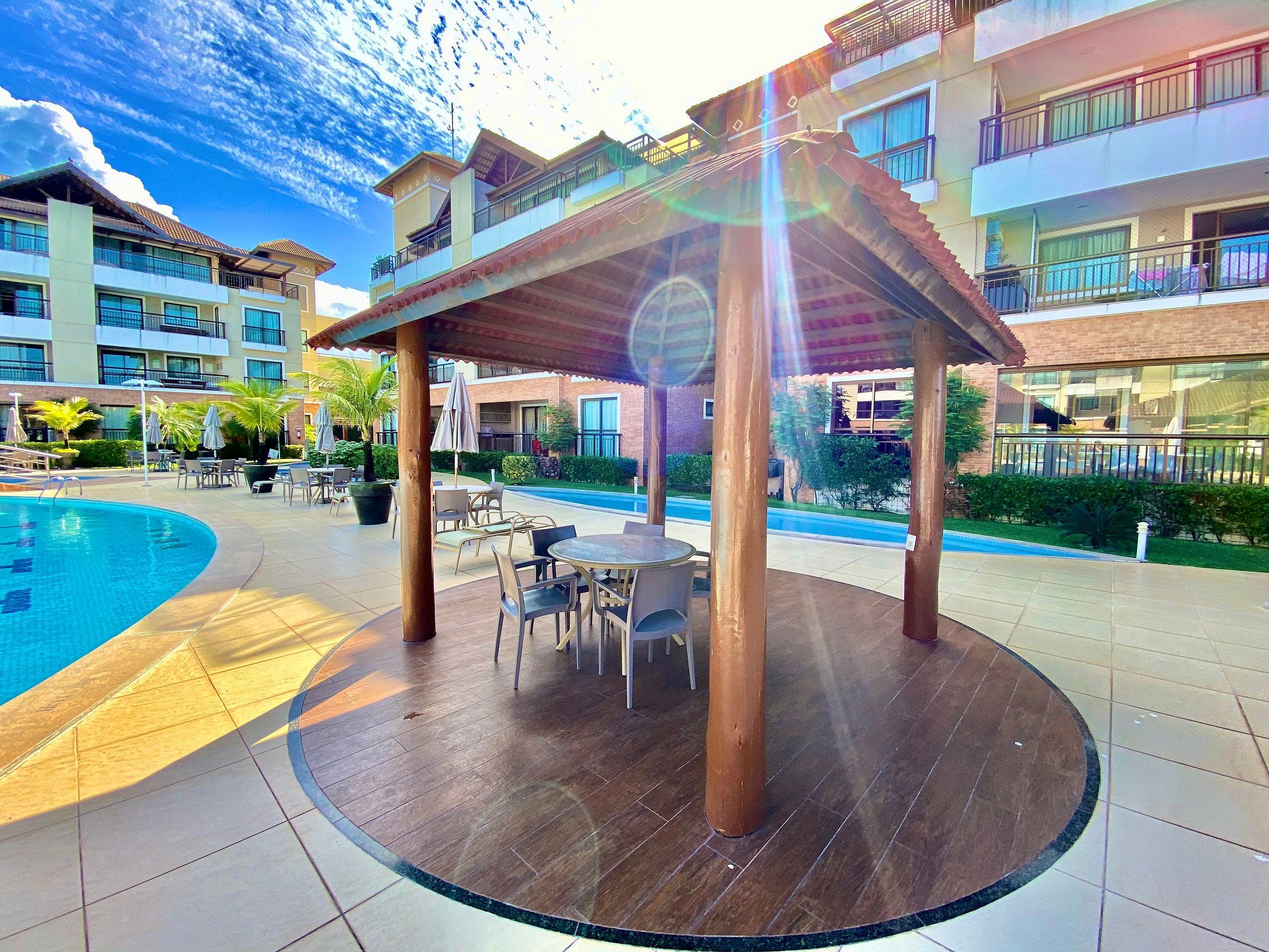 A poolside area with a covered seating area, outdoor tables, and chairs, surrounded by buildings with balconies.