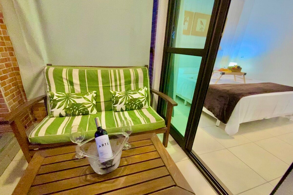 A green-striped sofa with a bottle of wine and glasses on a wooden table, a bed with a brown blanket, and a view through a glass door.