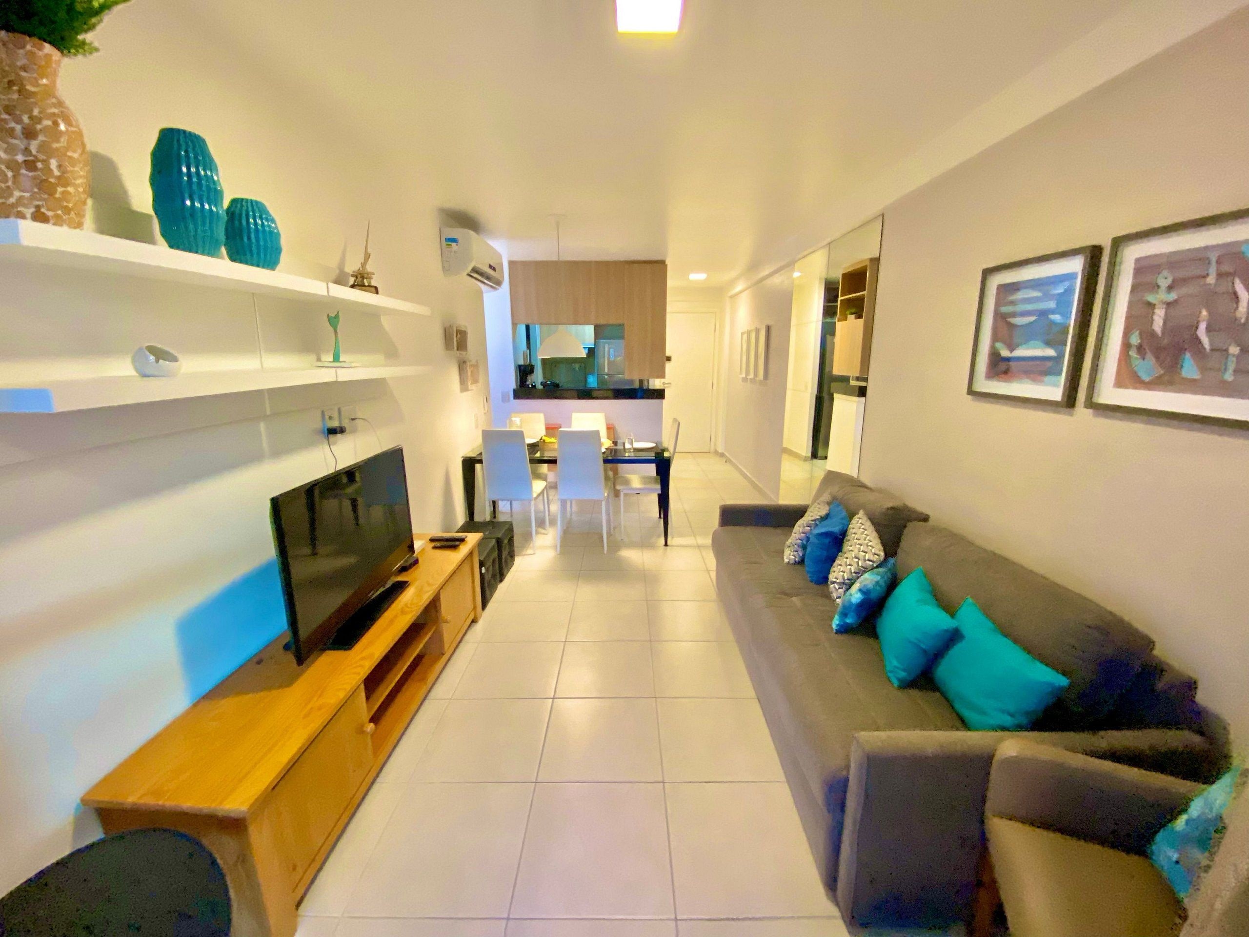 A modern living room with a flat-screen TV, a wooden entertainment unit, a grey sofa with blue cushions, and a dining area in the background.