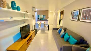 A modern living room with a flat-screen TV, a wooden entertainment unit, a grey sofa with blue cushions, and a dining area in the background.