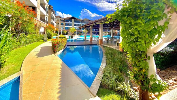 Una piscina con borde de baldosas, rodeada de vegetación y un edificio con varios balcones.
