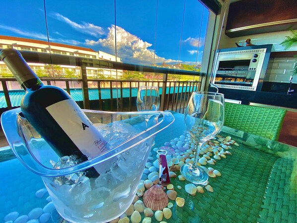 A glass bowl with a bottle and glasses on a table, surrounded by seashells, with a view of buildings and a clear sky.