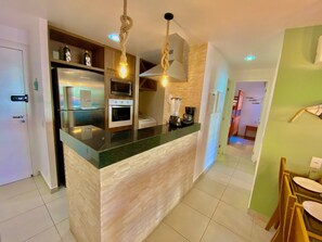 A modern kitchen with a central island, built-in appliances, and a dining area with chairs. - Condominio Mediterranee Residence por Be My Guest (Aquiraz)