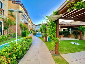 A pathway leading to a pool with surrounding greenery and a building with balconies. - Condominio Mediterranee Residence por Be My Guest (Aquiraz)