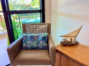 A chair with patterned cushions, a small wooden boat model, and a view of greenery through a window. - Condominio Mediterranee Residence por Be My Guest (Aquiraz)