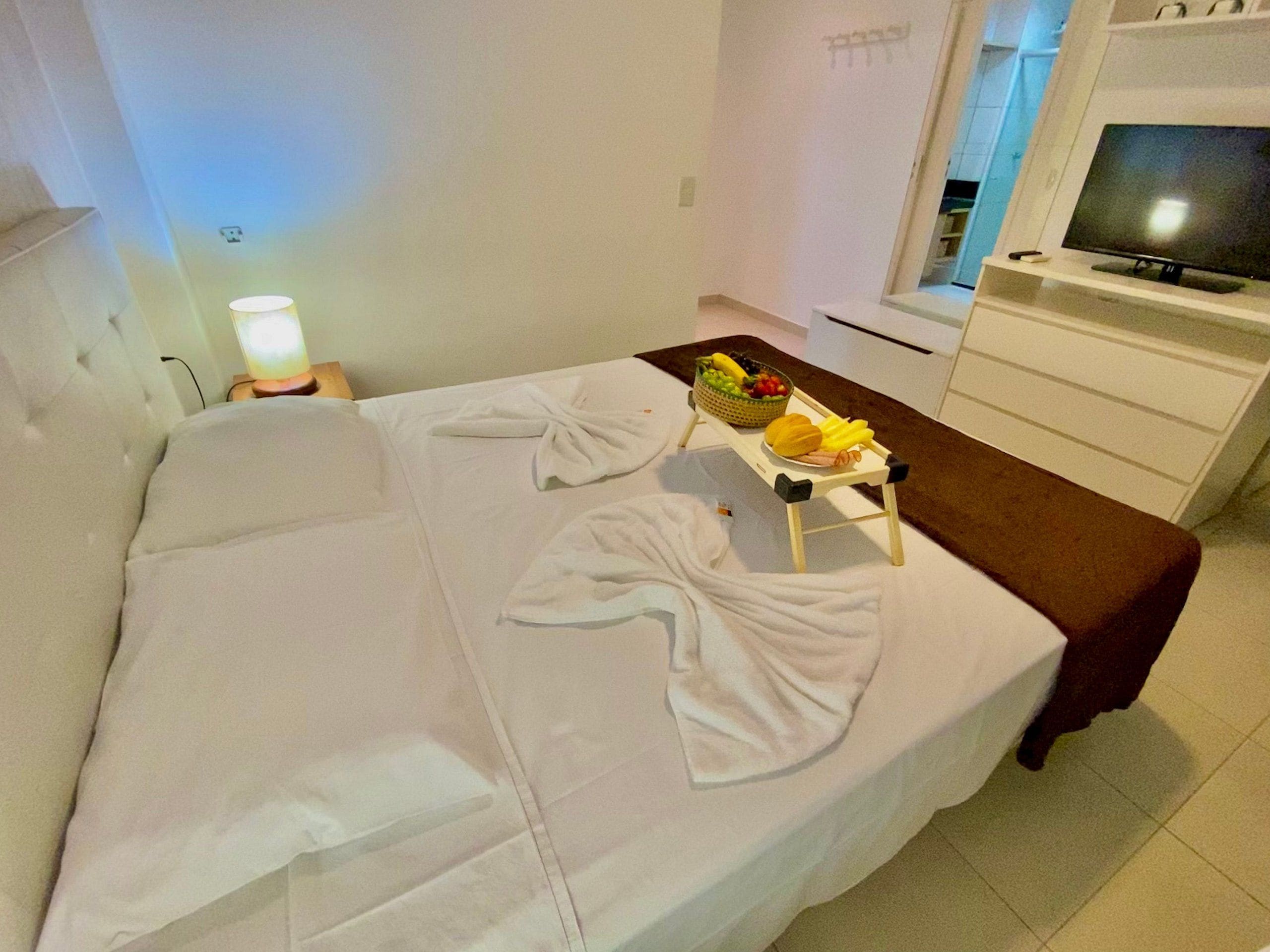 Quarto de hotel com cama, luminária de cabeceira, mesinha com frutas e televisão em um suporte.