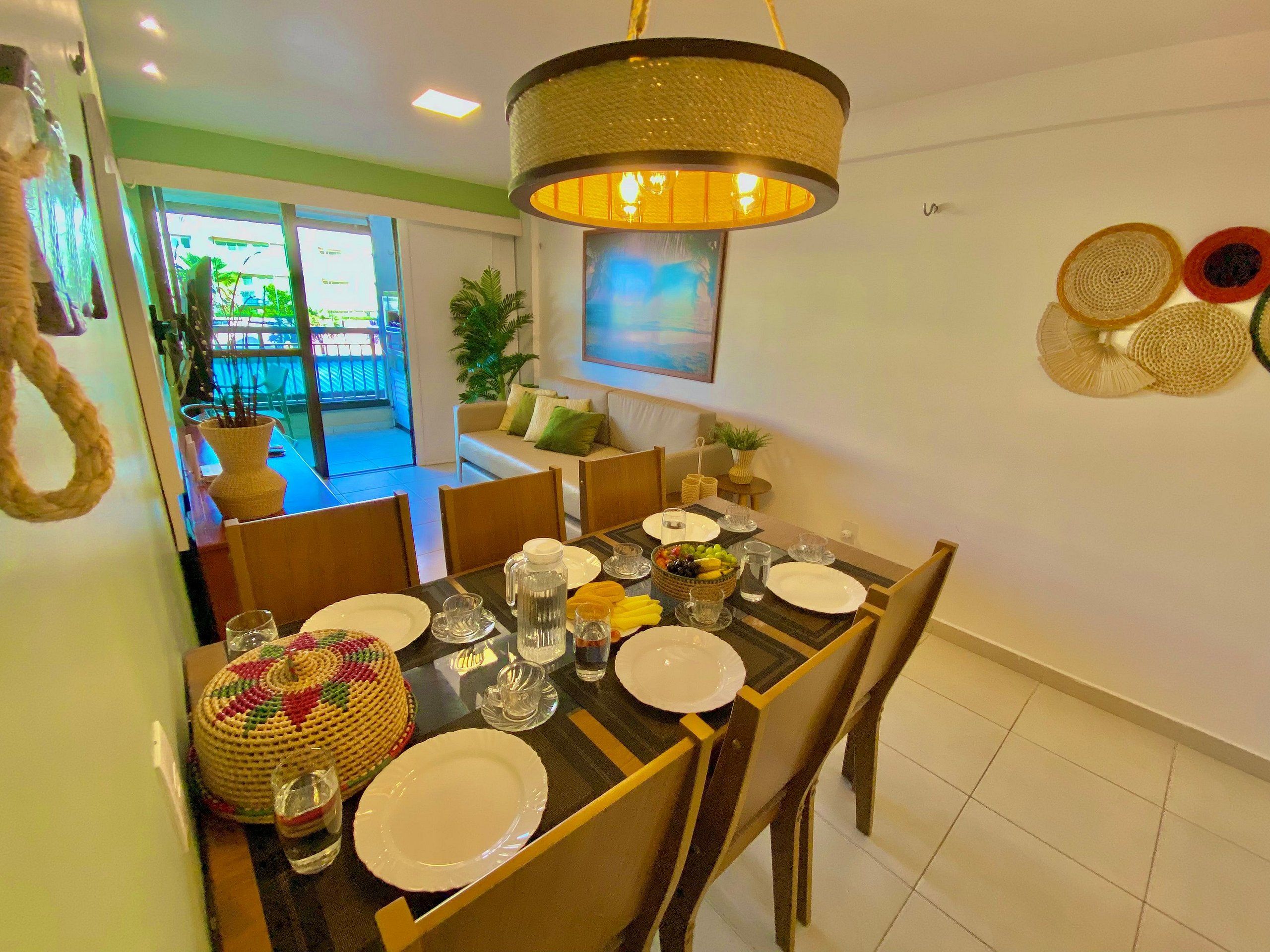 Uma área de refeições com mesa posta para quatro pessoas, cadeiras, luminária pendente e vista para a área externa.