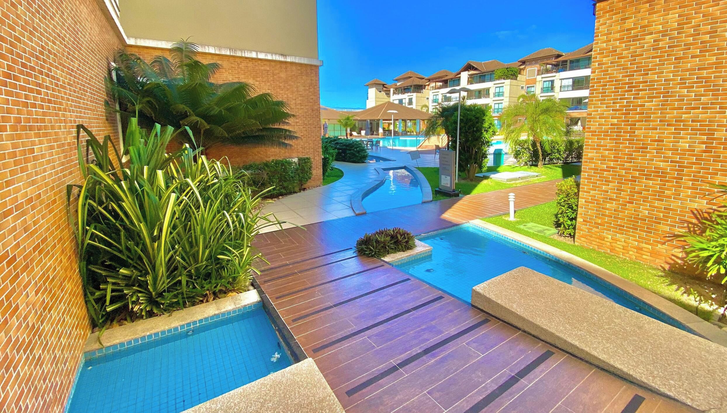 Uma área de piscina com muro de tijolos, plantas de palmeira e vista para um prédio de vários andares.