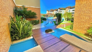 A swimming pool area with a brick wall, palm plants, and a view of a multi-story building. - Condominio Mediterranee Residence por Be My Guest (Aquiraz)
