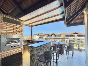 An outdoor kitchen area with a grill, bar counter, and seating. - Condominio Mediterranee Residence por Be My Guest (Aquiraz)
