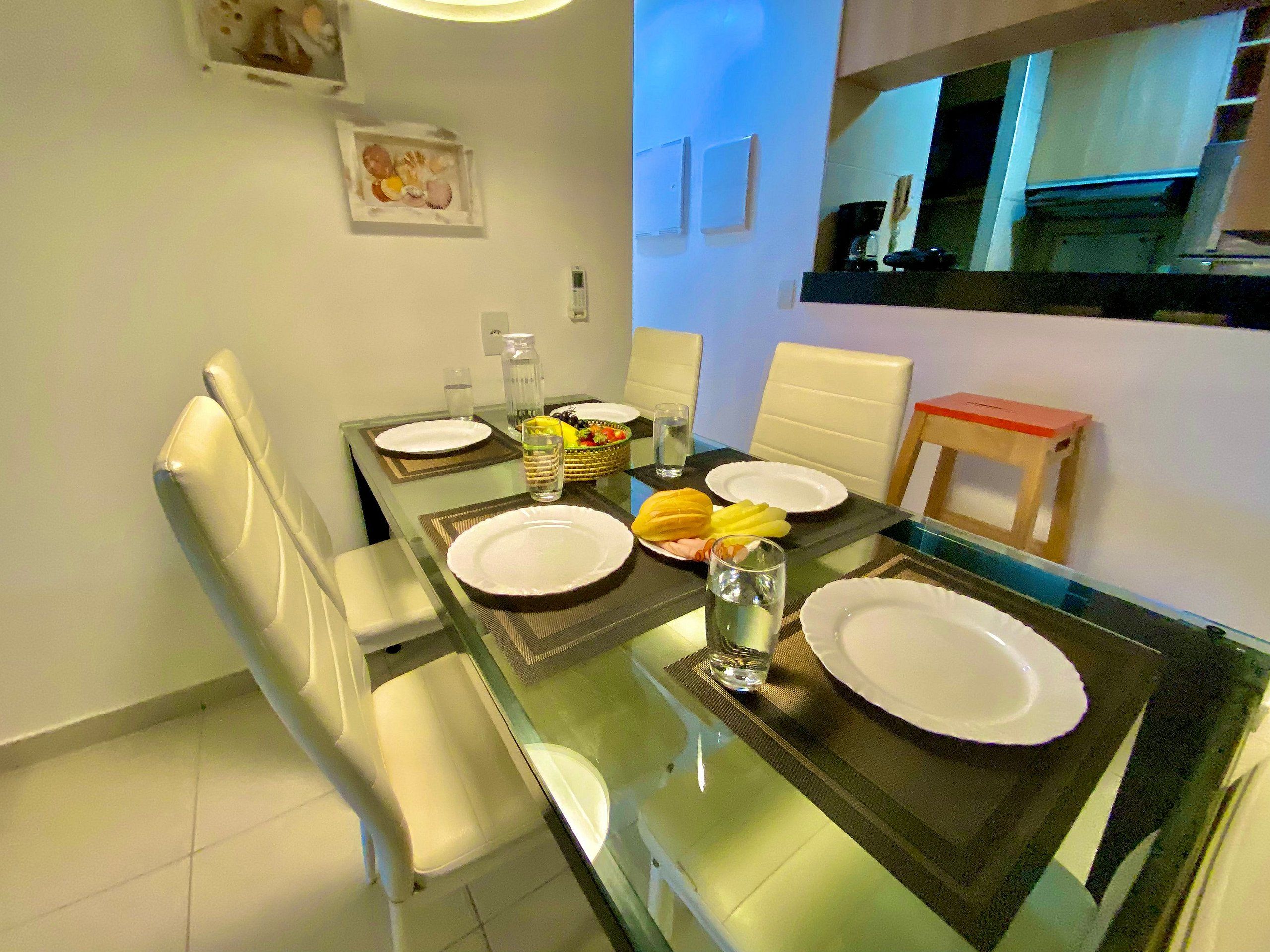 Uma sala de jantar com mesa de vidro, pratos brancos e uma cesta de frutas.
