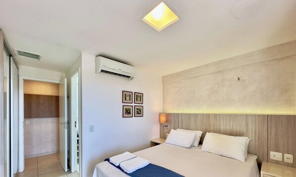 Una habitación de hotel con una cama, un equipo de aire acondicionado, una lámpara de noche y una puerta que da a un baño.