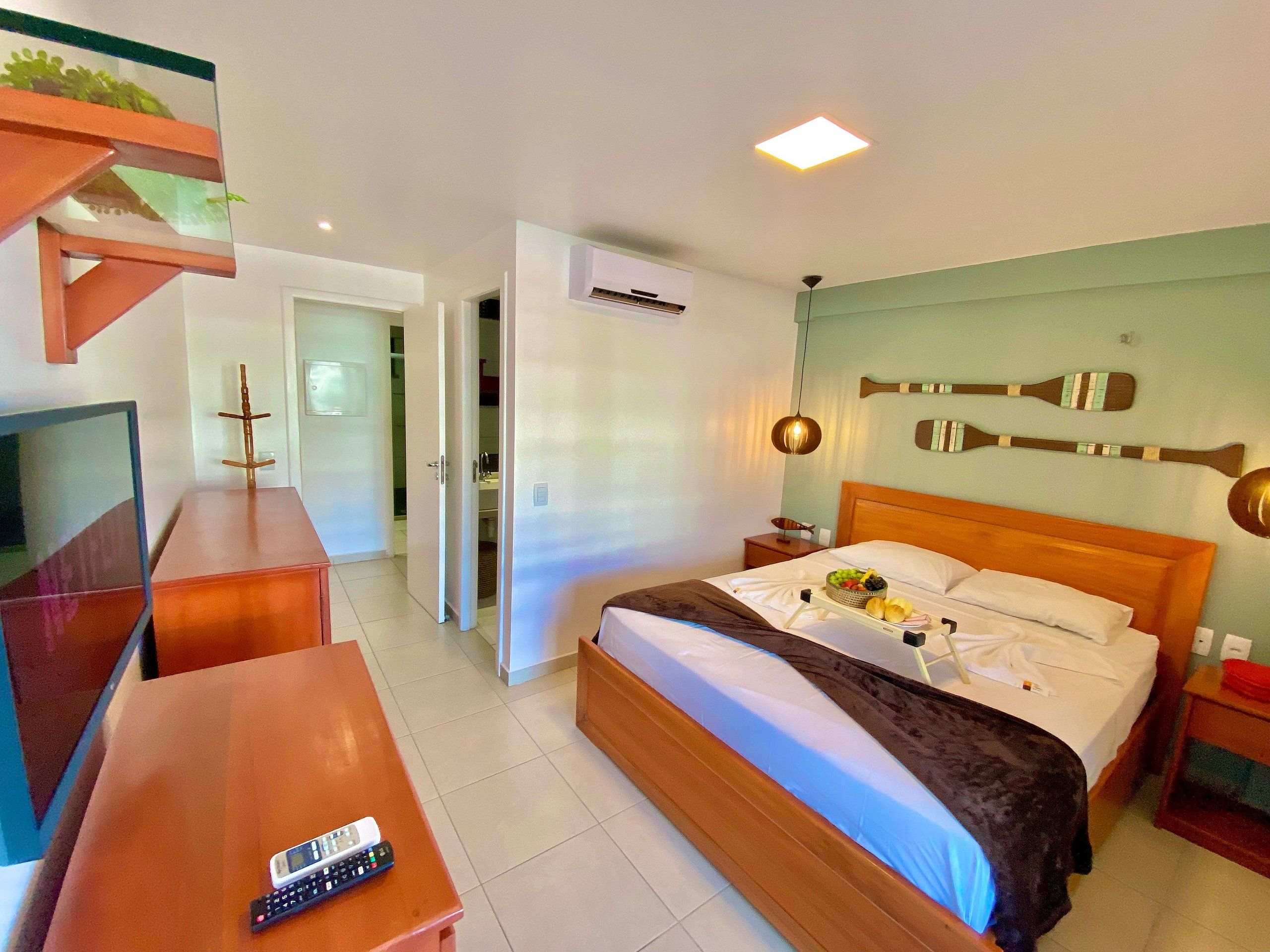 Quarto de hotel com cama, mesa de cabeceira, televisão e escrivaninha de madeira.