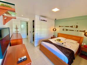 Quarto de hotel com cama, mesa de cabeceira, televisão e escrivaninha de madeira.
