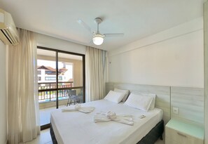A hotel room with a bed, a ceiling fan, a window with curtains, and a view of buildings outside. - Condominio Mediterranee Residence por Be My Guest (Aquiraz)