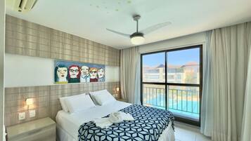Habitación de hotel con cama, ventilador de techo, ventana grande con cortinas, un cuadro en la pared y vista a un edificio y una piscina al aire libre.