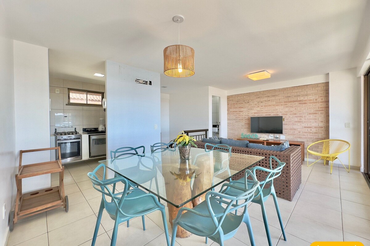 A modern kitchen with stainless steel appliances, a dining area with a glass table and blue chairs, a brick accent wall, and a TV mounted on the wall.