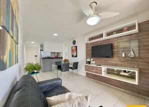 A modern living room with a black sofa, a dining area, a kitchen in the background, and a mounted TV on a wooden wall.