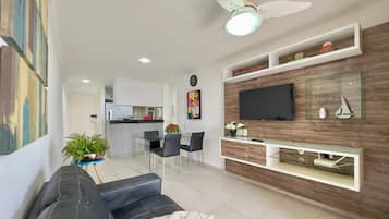Uma sala de estar moderna com um sofá preto, uma área de jantar, cozinha ao fundo e uma televisão fixada numa parede de madeira.