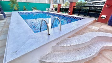 Una piscina con un patrón de baldosas de ajedrez, dos escaleras metálicas y una pared verde al fondo.