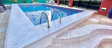 A swimming pool with a checkered tile pattern, two metal ladders, and a green wall in the background.