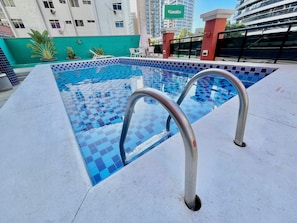Una piscina con baldosas de diseño ajedrezado, barandillas metálicas a su alrededor y un muro verde al fondo.