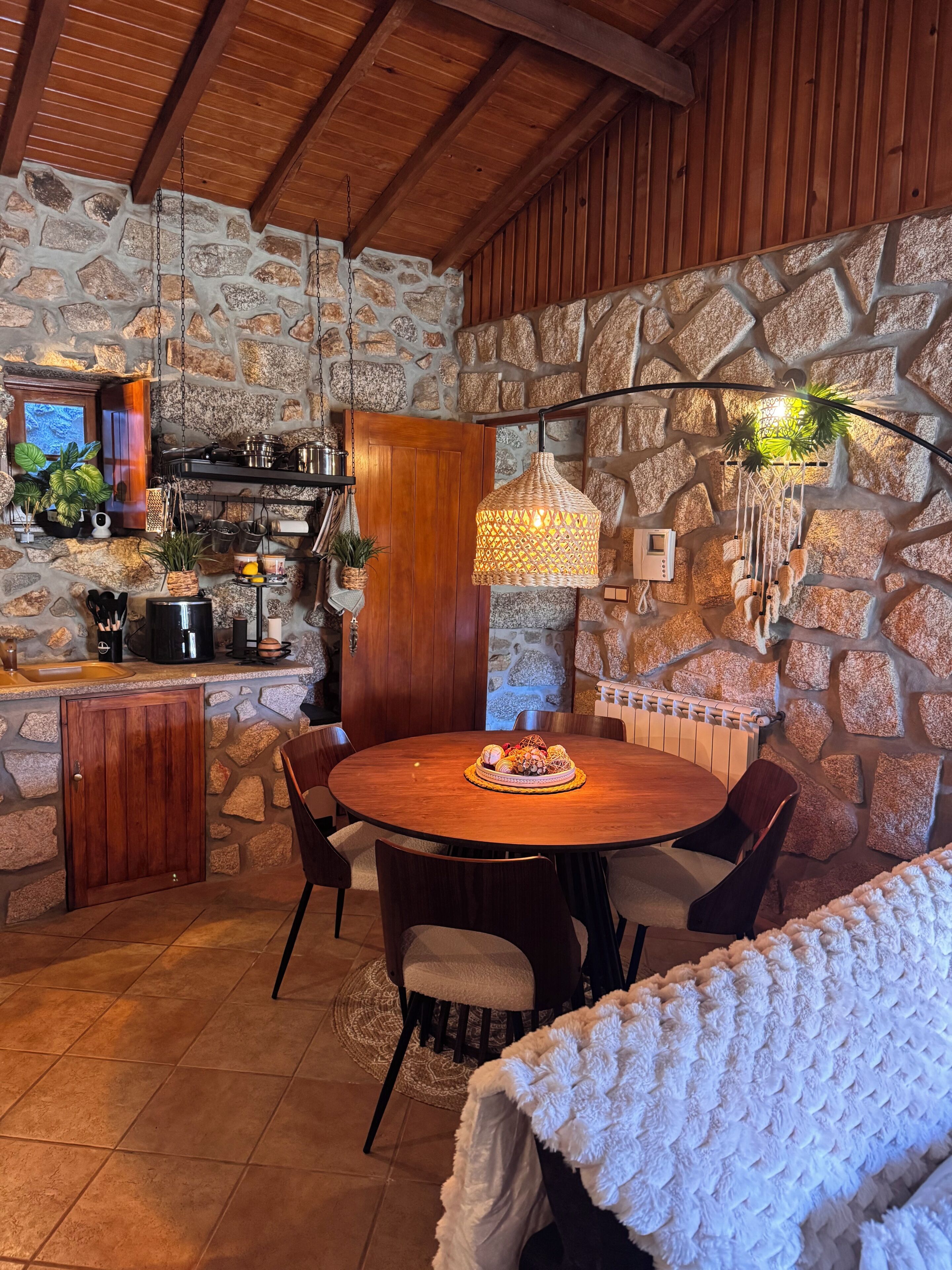 Une cuisine aux murs de pierre et au plafond en bois, une table à manger ronde entourée de chaises, et un luminaire suspendu.