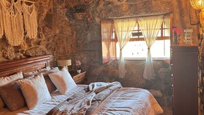 A cozy room with a stone wall, a bed with a macrame headboard, a wooden dresser, and a window with curtains.
