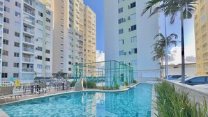A swimming pool with tiled flooring, surrounded by tall residential buildings, and a green metal structure in the pool area. - Mirante Condominio Clube (Fortaleza)