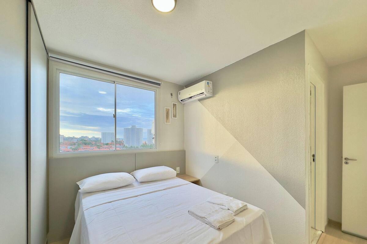 A hotel room with a bed, a window with a city view, an air conditioning unit, and a door.