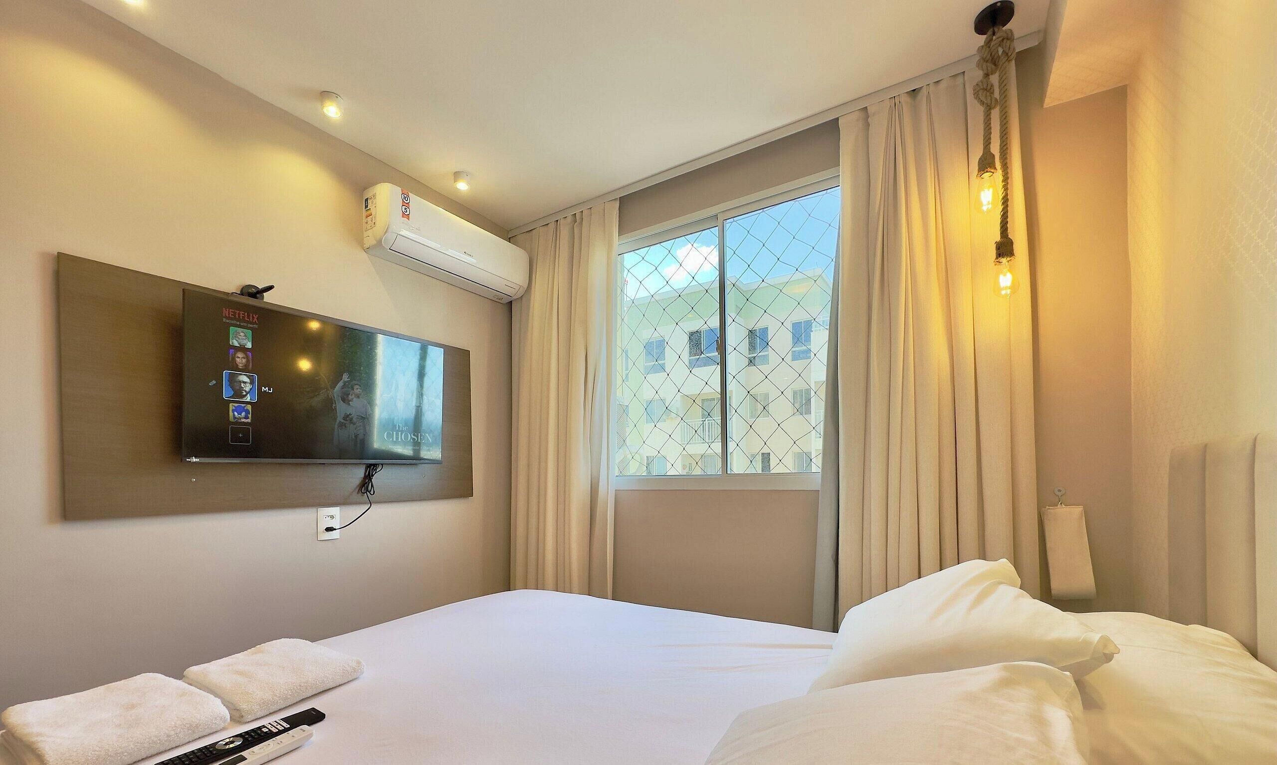 Een hotelkamer met een bed, een aan de muur bevestigde televisie, een raam met gordijnen en een airconditioning.