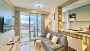 A modern living room with a sofa, a TV, a balcony with a view of buildings, and a kitchen area. - Mirante Condominio Clube (Fortaleza)