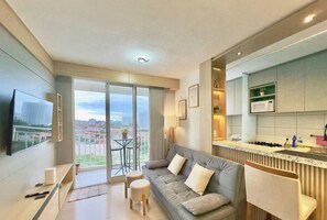 Un salon moderne comprenant un canapé, un téléviseur, un balcon avec vue sur les immeubles et un coin cuisine.