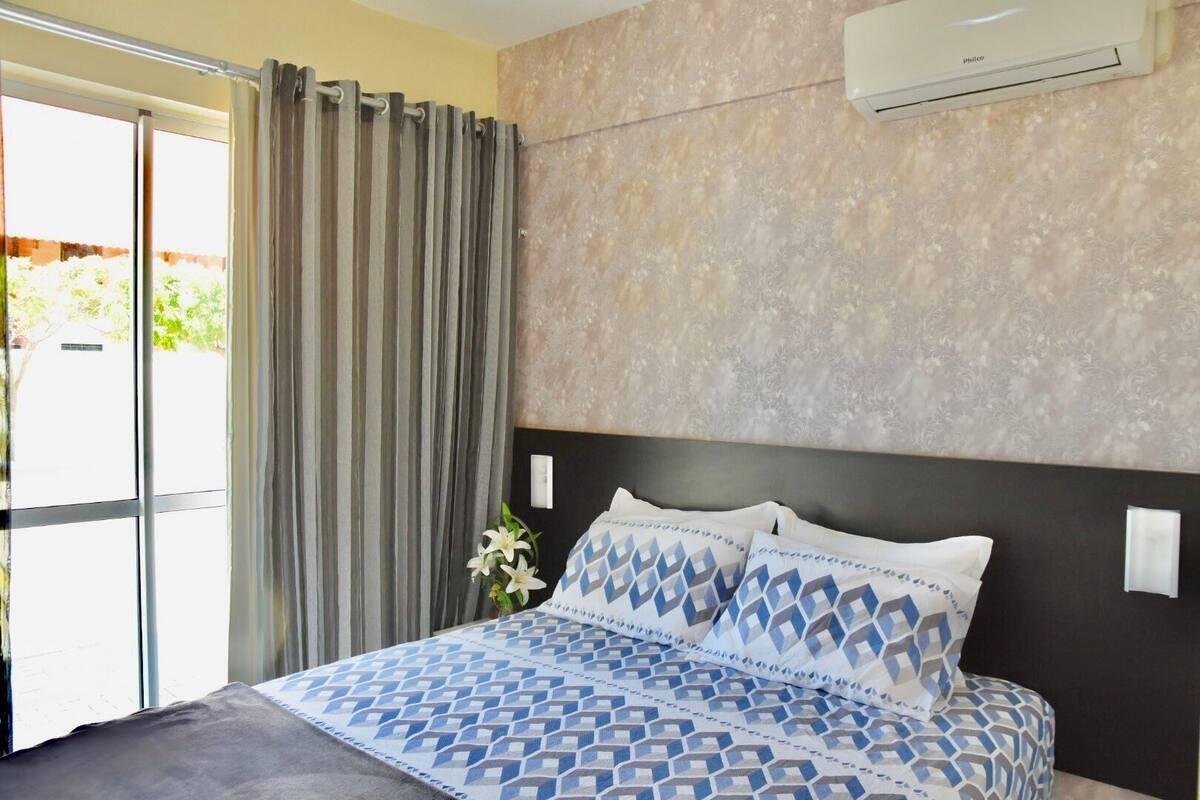 A bedroom with a bed, curtains, an air conditioner, and a view of the outdoors.
