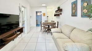 En stue med fjernsyn, sofa, spisebord med stole og et køkken i baggrunden.