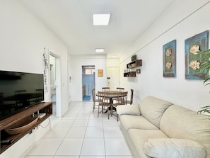 A living room with a TV, a sofa, a dining table with chairs, and a kitchen in the background. - Palm Beach Porto das Dunas - CE (Aquiraz)