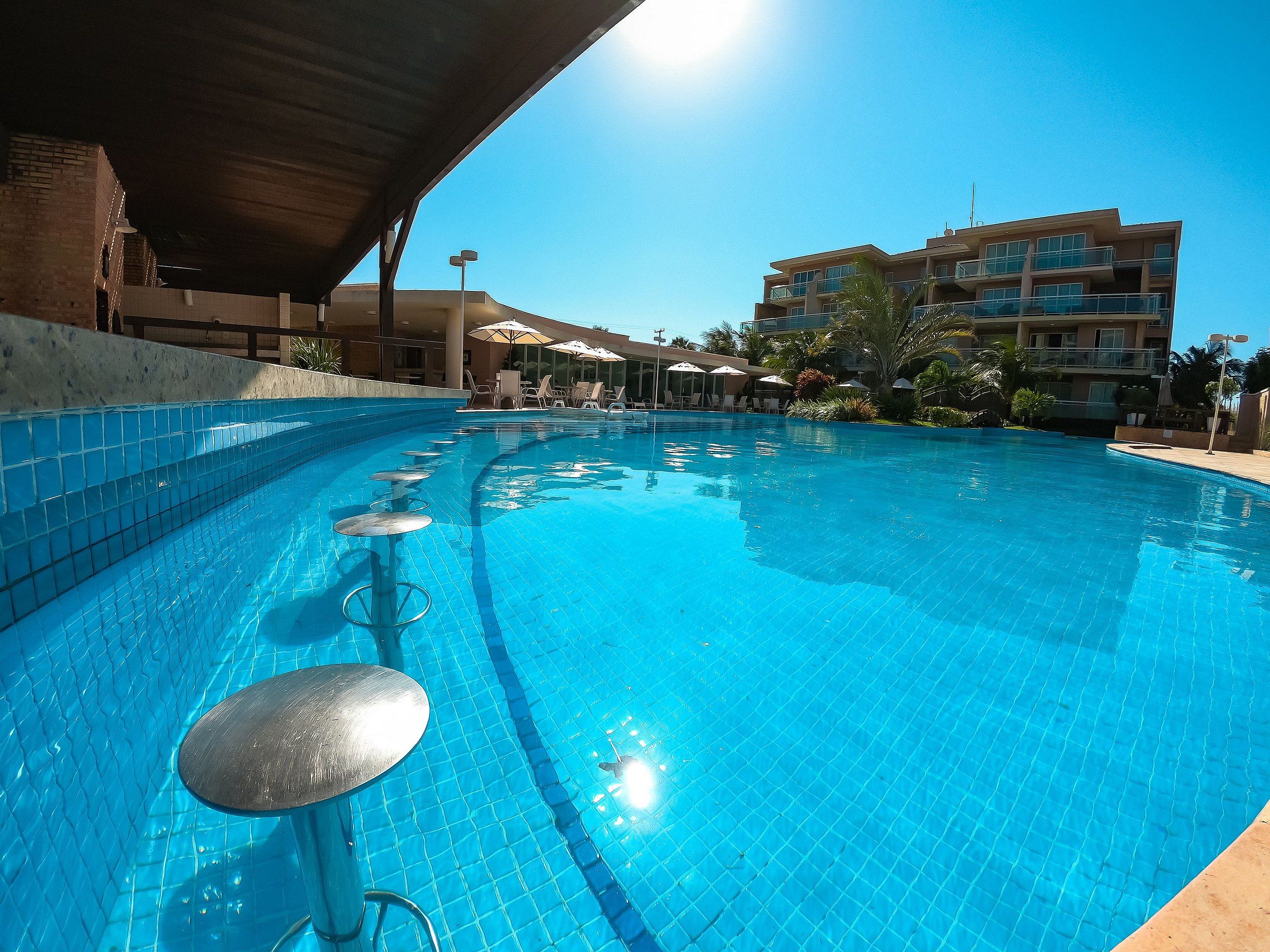 Piscina com borda azulejada, espreguiçadeiras, guarda-sóis e um edifício ao fundo.