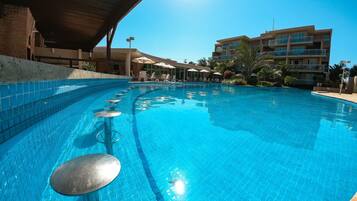 Piscina com borda azulejada, espreguiçadeiras, guarda-sóis e um edifício ao fundo.