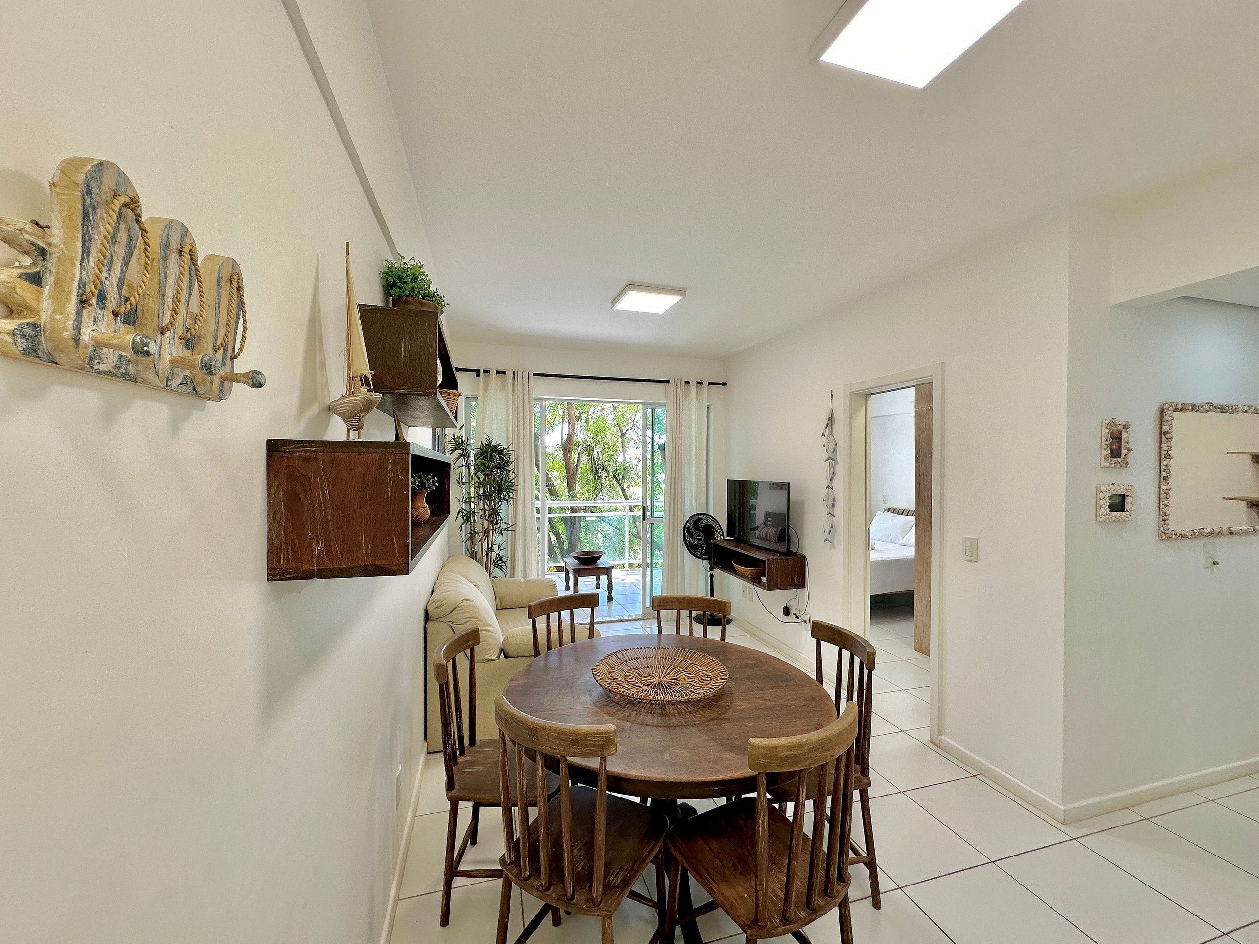 A dining area with a round wooden table, six chairs, a TV mounted on the wall, and a large window with curtains.