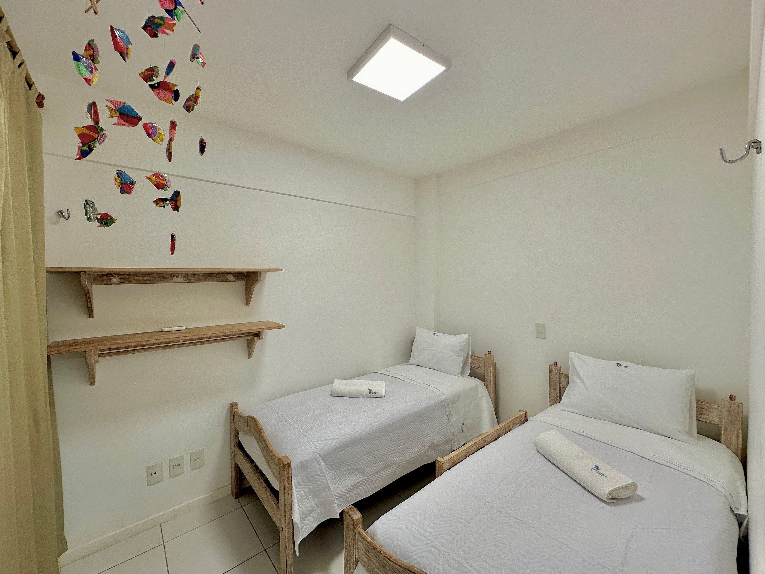 Une pièce avec deux lits en bois, du linge de lit blanc, un luminaire au plafond et des étagères fixées au mur.